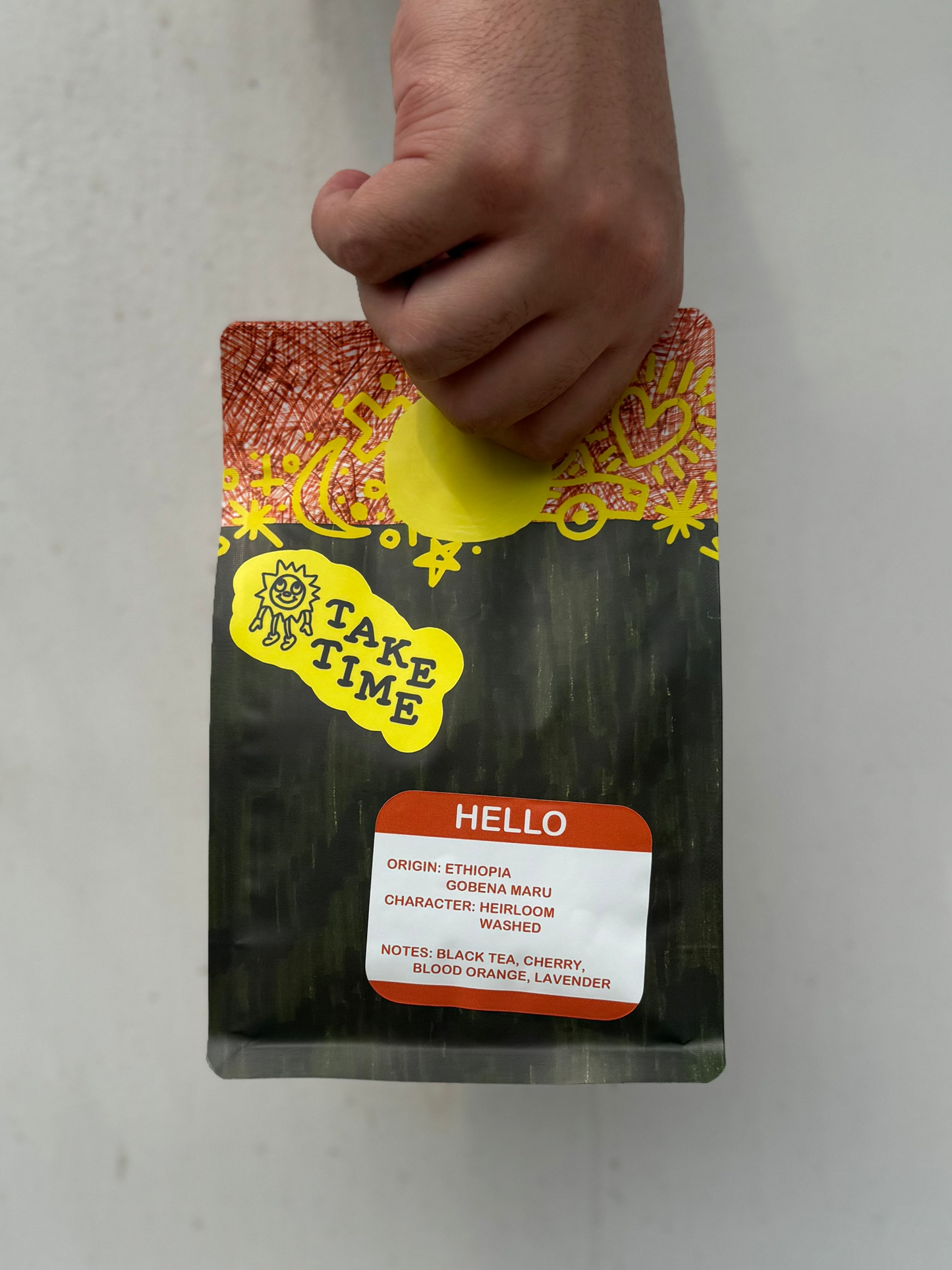 A hand holding a bag of Take Time Hello herbal tea against a plain light gray background. The bag is black with a red and yellow patterned top, a yellow Take Time logo, and a white label with red text listing the tea notes.