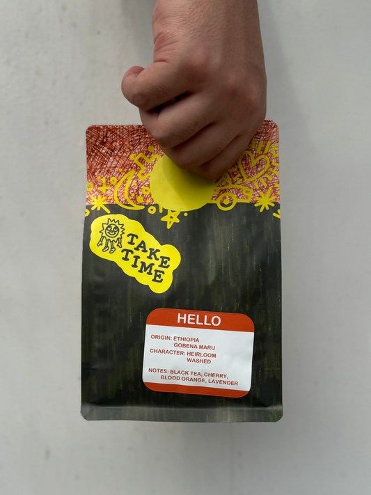 A hand holding a bag of Take Time Hello herbal tea against a plain light gray background. The bag is black with a red and yellow patterned top, a yellow Take Time logo, and a white label with red text listing the tea notes.